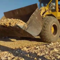 Gelbe Planierraupe vor blauem Himmel in Syrien. Zwei Männer sitzen in der Fahrerkabine. Die Raupe fährt auf ockerbrauner Erde. In der breiten Schaufel ist auch Erde.