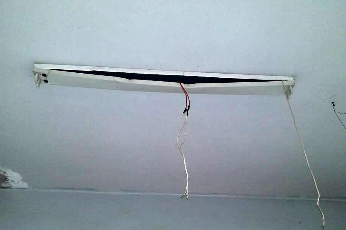 Eine verbogene Lampenhalterung an der Decke, aus der Kabel herunterländen. Die Leuchtstoffröhren fehlen.