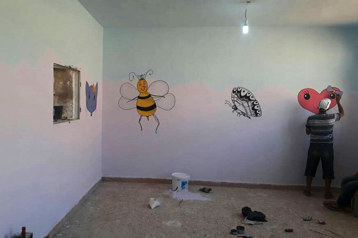 Ein Mann bemalt die renovierten Wände eines Klassenzimmers mit bunten Figuren.