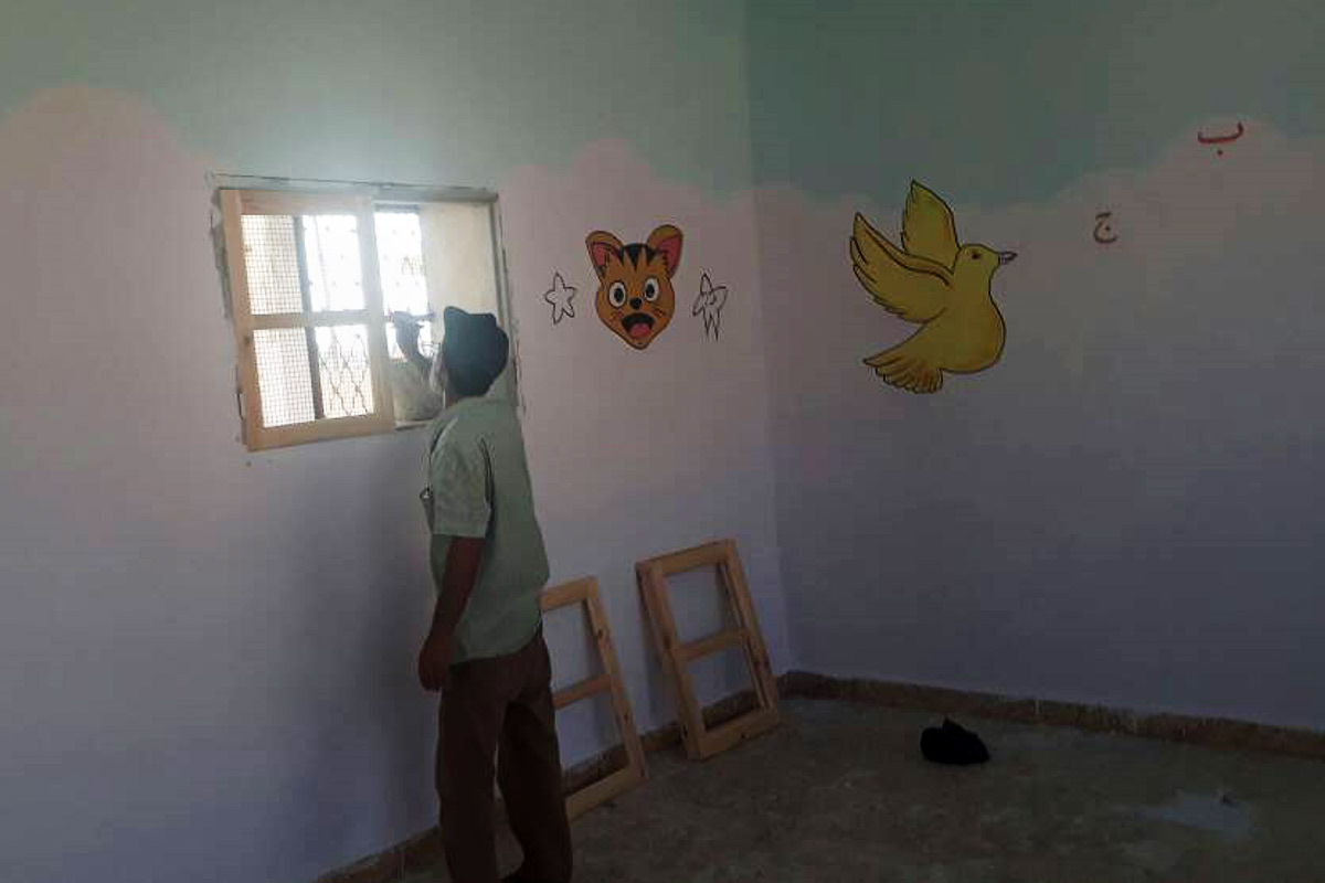 Ein Mann bringt neue Fensterläden aus Holz in einem Klassenzimmer an. An den Wänden sind Bilder von einer Katze und einem gelben Vogel.