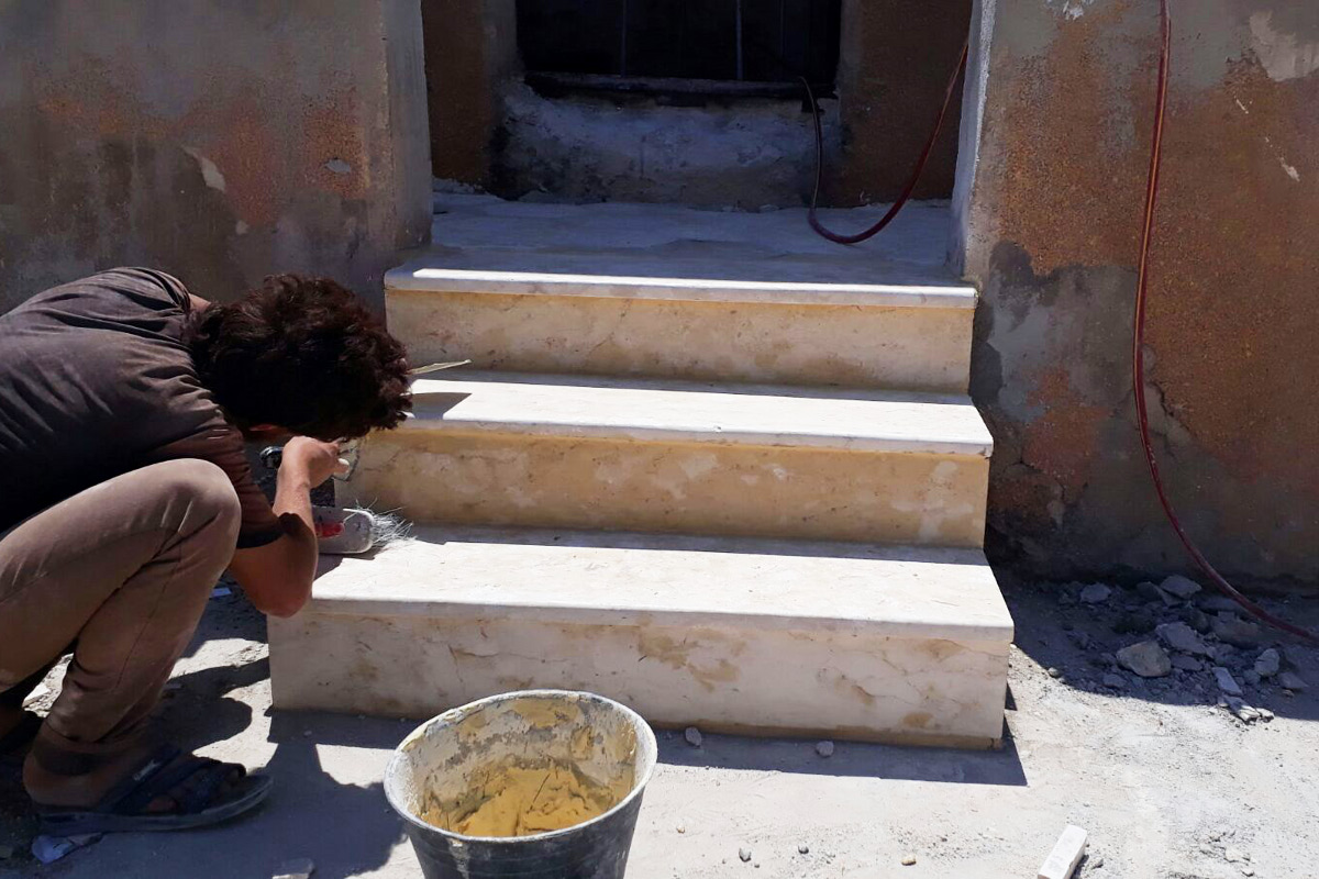 Ein Mann repariert die Treppenstufen am Angang der Schule. Im Vordergrund steht ein Eimer mit Mörtel.