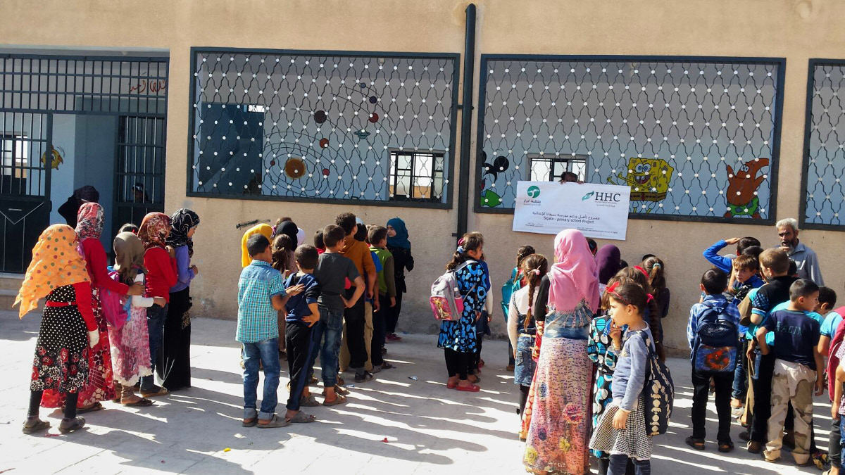 Kinder stehen in vier Reihen vor einem braunen Schulgebäude mit großen Fenstern. Rechts sieht man einen älteren Mann mit Bart. Die Kinder tragen bunte Kleidung. Einige Mädchen tragen Kopftücher.