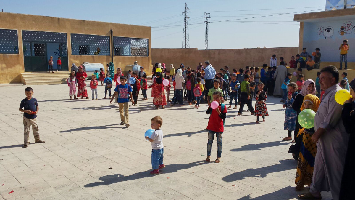Kinder spielen draußen im Hof einer Schule. Einige haben bunten Luftballons. Man sieht auch einige Erwachsene. Hinten sieht man das renovierte braune Schulgebäude.