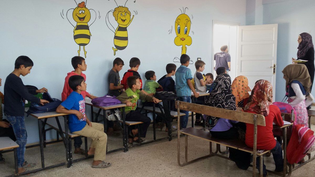 Eine Gruppe Mädchen und Jungen sitzt in einem Klassenraum. Rechts steht eine Frau mit Kopftuch. Auf die Wand sind Cartoonfiguren gemalt: Bienen und ein gelber Vogel.