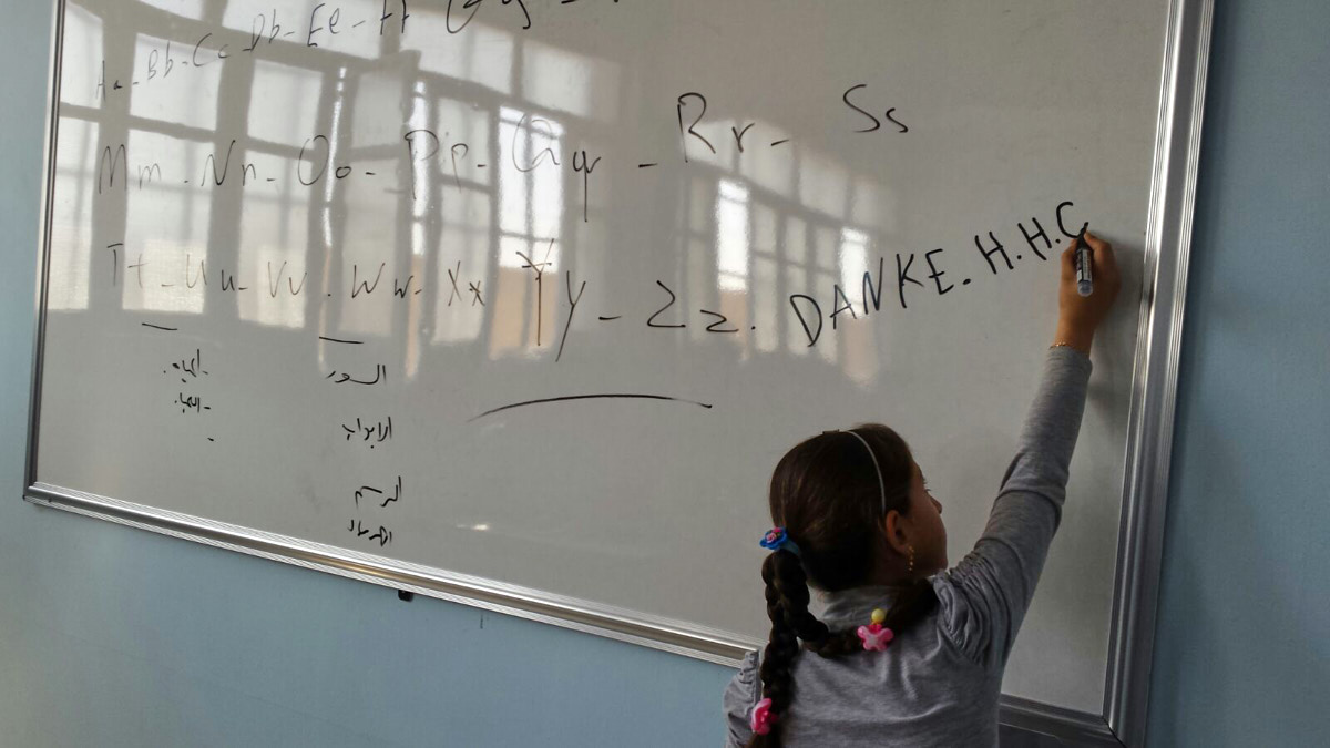 Ein Mädchen schreibt "DANKE HHC" an eine weiße Tafel. Die hellen Fenster des Klassenraums spiegeln sich in der Tafel.