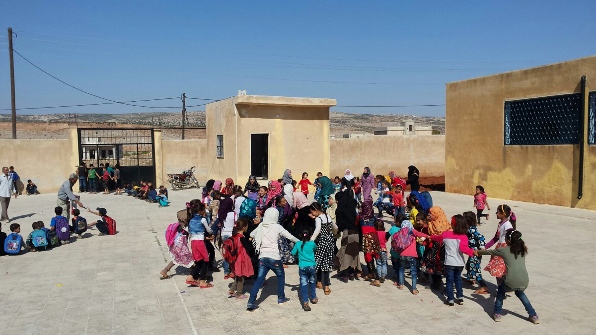 Kinder spielen draußen im Hof einer Schule. Sie fassen sich an den Händen. Hinten sieht man zwei grau-braune Gebäude und ein großes Tor.