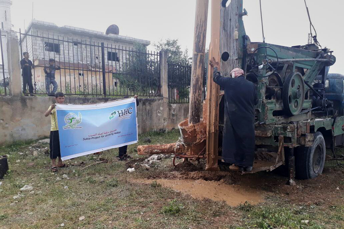 Ein Mann steht hinten auf einem großen Brunnenbohrfahrzeug. Daneben halten zwei Jungen ein Schild mit dem HHC-Logo hoch.