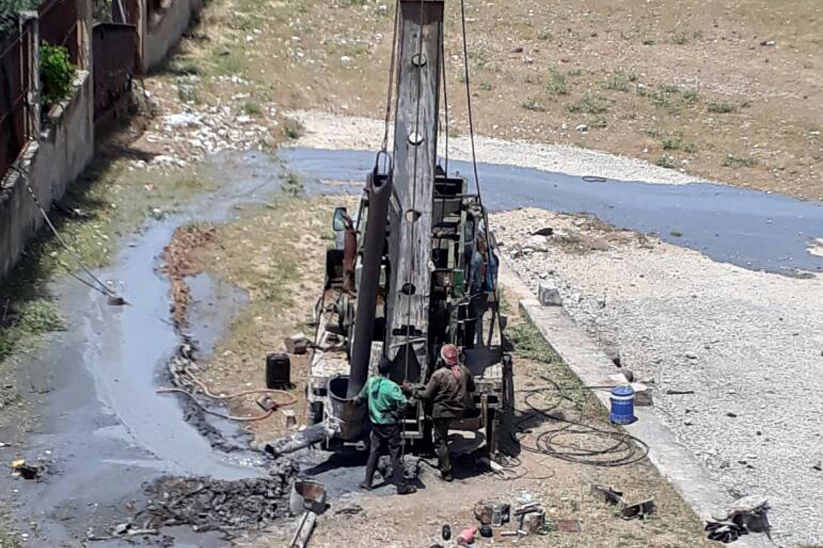Zwei Männer stehen an einem Brunnenbohrgerät. Wasser sprudelt aus dem Boden und fließt in den Sand.