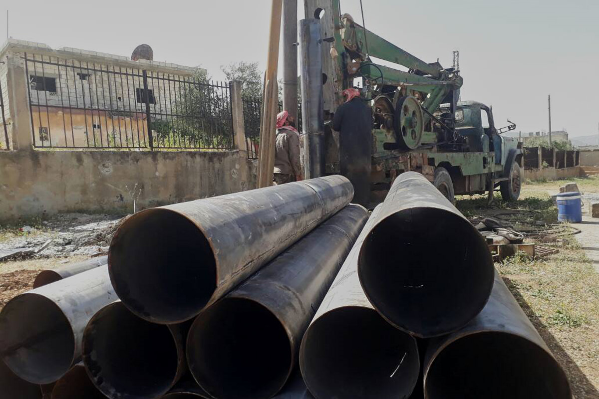 Große Rohrleitungen aus Stahl liegen auf einem Stapel. Dahinter sieht man ein Brunnenbohrgerät.