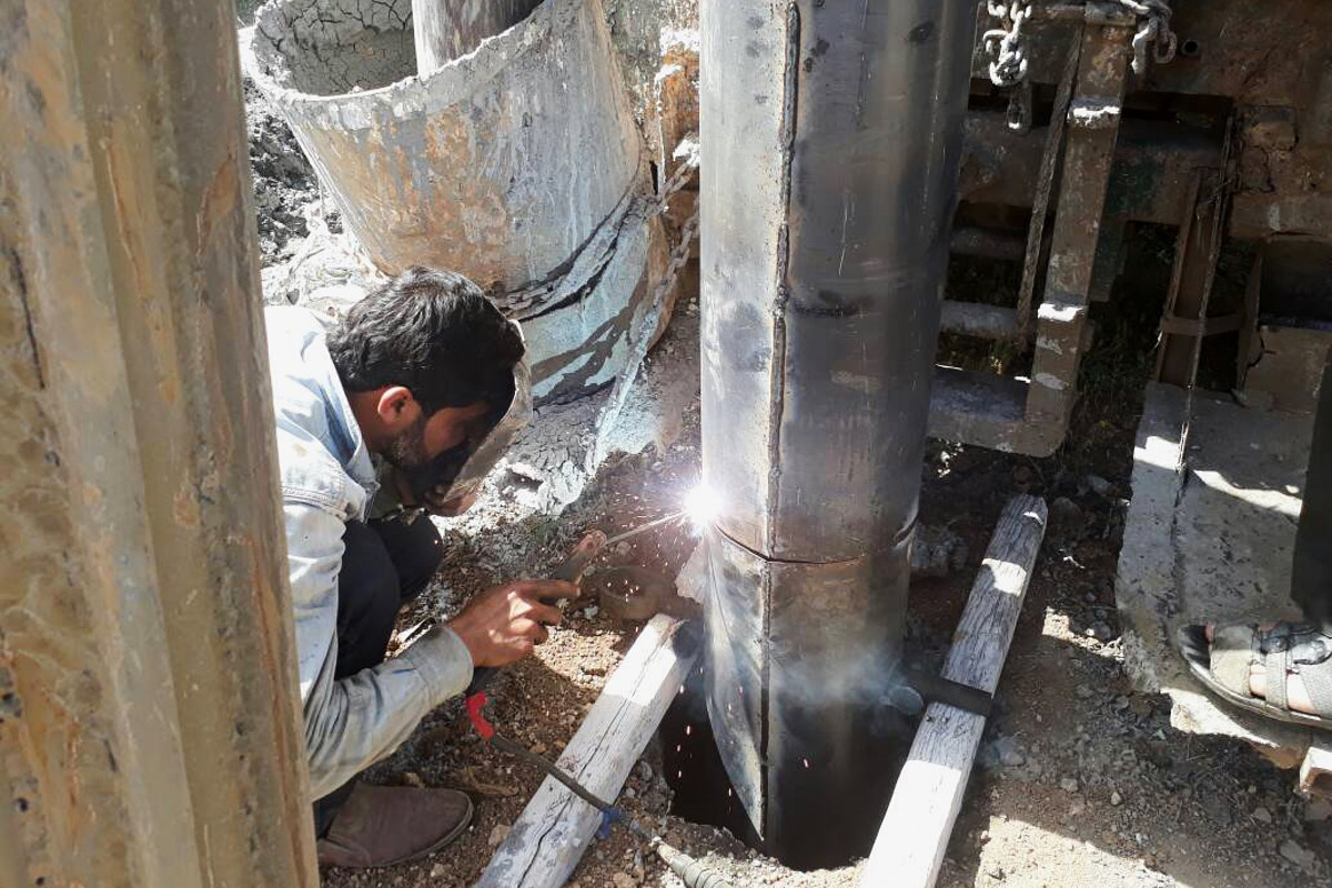 Ein Mann hockt auf dem Boden und schweißt eine dicke Rohrleitung, die aus der Erde ragt.